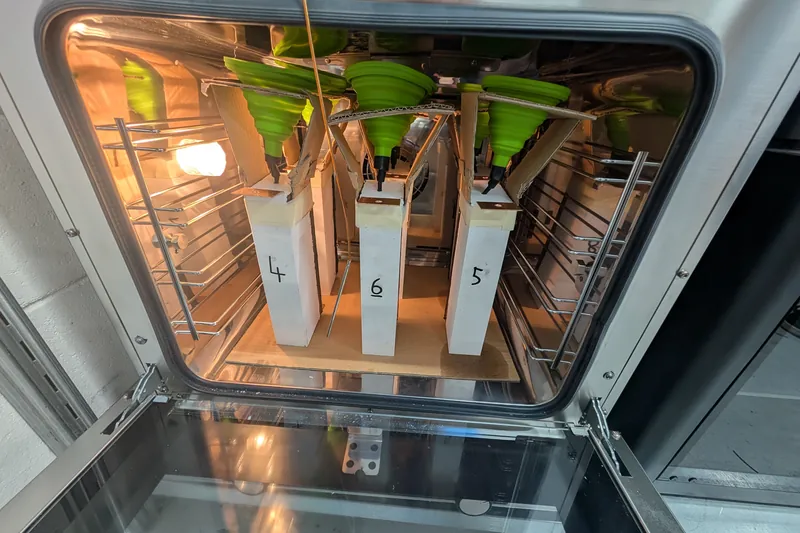Three numbered white modules inside an open laboratory oven, each fed by a green silicone funnel.